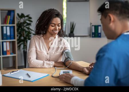 Adult asian man doctor measures pressure of smiling young black female patient in clinic office interior Stock Photo