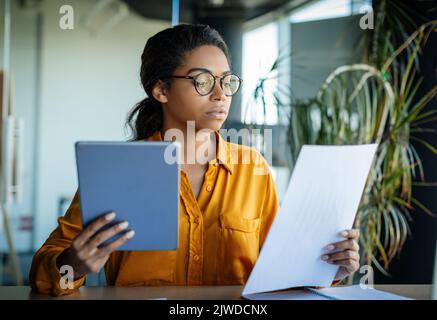 Serious black woman entrepreneur using digital tablet and reading documents, working through financial papers Stock Photo