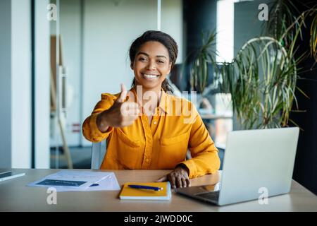 Happy african american female office manager sitting at table with laptop, showing thumb up and smiling in office Stock Photo