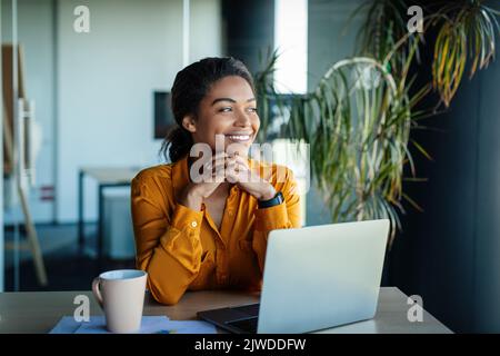 Portrait of dreamy african american businesswoman sitting at desk in office and thinking while working on laptop Stock Photo