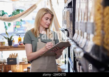 Female Owner Of Sustainable Plastic Free Grocery Store Checking Stock On Shelves Stock Photo