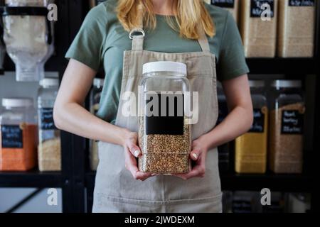 Close Up Of Sales Assistant In Sustainable Plastic Free Wholefood Store Holding Container With Blank Label Stock Photo