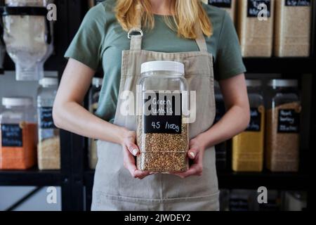 Close Up Of Sales Assistant In Sustainable Plastic Free Wholefood Store Holding Container Of Green Lentils Stock Photo