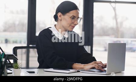smiling businesswoman in eyeglasses and formal wear typing on laptop near smartphone,stock image Stock Photo