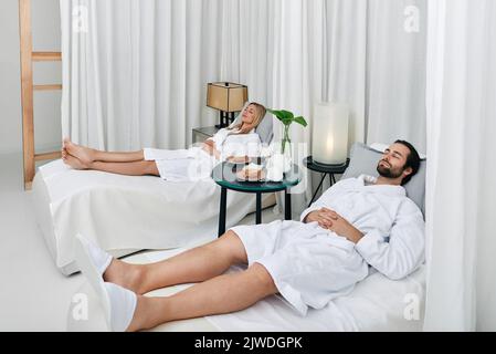 Relaxed beautiful couple with eyes closed laying down on white beds, resting in lounge zone of wellness center after spa procedures Stock Photo