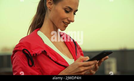 Positive brunette sportswoman using mobile phone on blurred roof in evening,stock image Stock Photo