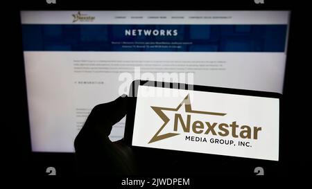 Person holding cellphone with logo of American television company Nexstar Media Group Inc. on screen in front of webpage. Focus on phone display.