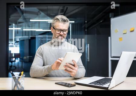 Senior experienced gray haired man in glasses using tablet computer, businessman at work inside modern office building Stock Photo