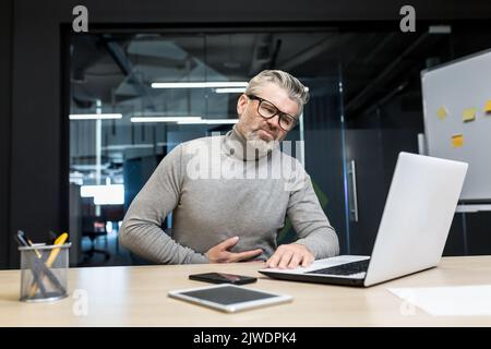 Sick gray-haired businessman poisoned, boss holding stomach, having severe stomach pain mature man working in modern office using laptop. Stock Photo