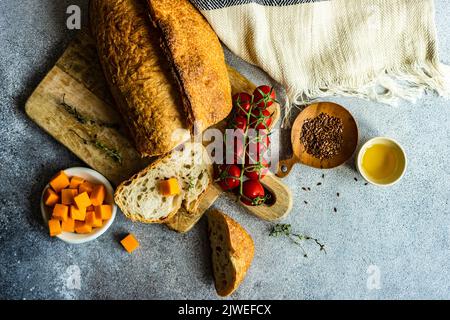 Overhead view of a loaf of sourdough bread on a chopping board with cherry vine tomatoes, cubes of cheese, olive oil and flax seeds Stock Photo