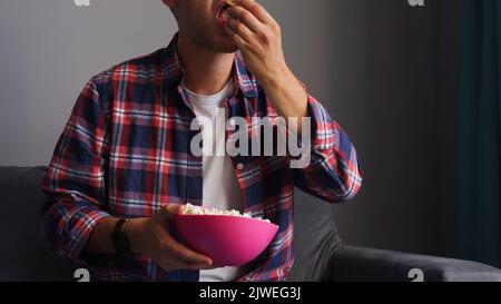 A young man watches a movie and eats roasted corn while sitting in the living room on the couch in his home. Stock Photo