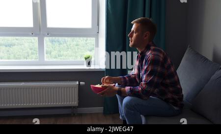 A young man watches a movie and eats roasted corn while sitting in the living room on the couch in his home. Stock Photo