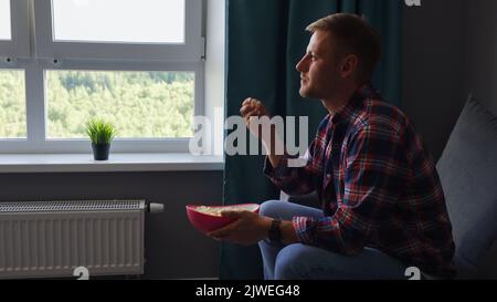 A young man watches a movie and eats roasted corn while sitting in the living room on the couch in his home. Stock Photo