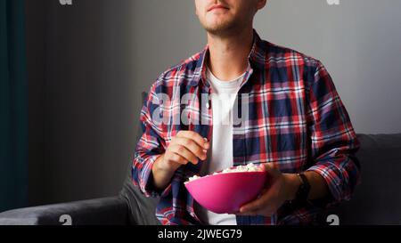 A young man watches a movie and eats roasted corn while sitting in the living room on the couch in his home. Stock Photo