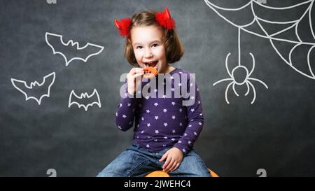 Kid in Halloween costume horns having fun, adorable happy child smiles and eats Halloween pumpkin cookie, studio portrait of cute little girl with dra Stock Photo