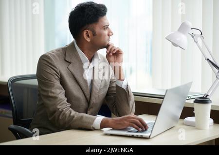 Portrait of pensive dreamy entrepreneur sitting at office desk with opened laptop and looking through window Stock Photo