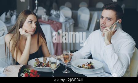 Busy young man is talking on mobile phone while his pretty girlfriend is feeling bored sitting at table in restaurant. Modern technologies, relationship and boredom concept. Stock Photo