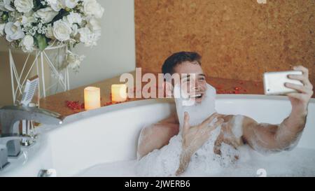 Cheerful young man with foam on beard is taking selfie using smartphone in hot tub in modern spa salon. He is laughing, gesturing, posing and looking at camera. Stock Photo