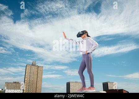 Woman outside wearing virtual reality headset with her arm outstretched, urban backdrop. Stock Photo