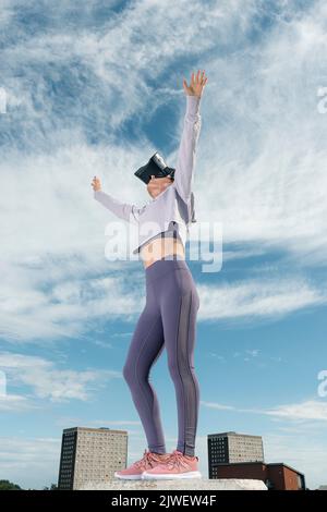 Woman outside wearing virtual reality headset with her arms outstretched, urban backdrop. Stock Photo