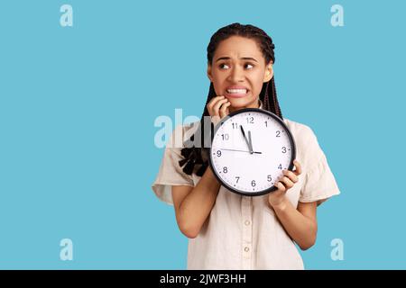 Portrait of impatient nervous woman with black dreadlocks biting her nails and holding big wall clock, deadline, wearing white shirt. Indoor studio shot isolated on blue background. Stock Photo