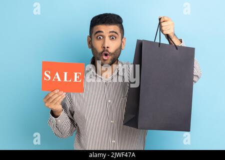Surprised man posing with shopping bags and card with sale inscription in hands, looking at camera with shocked expression, wearing striped shirt. Indoor studio shot isolated on blue background. Stock Photo