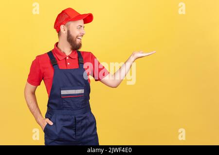 Portrait of smiling worker man standing and presenting copy space for advertisement on his palm, looking away, wearing overalls and red cap Indoor studio shot isolated on yellow background. Stock Photo