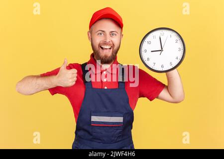 Portrait of happy smiling handyman standing with big wall clock in his hands, looking at camera and showing thumb up, wearing overalls and red cap. Indoor studio shot isolated on yellow background. Stock Photo