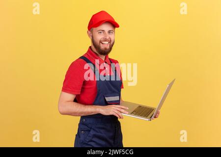 Side view of worker man wearing blue overalls, red T-shirt and cap working on laptop, expressing positive emotions, looking at camera. Indoor studio shot isolated on yellow background. Stock Photo