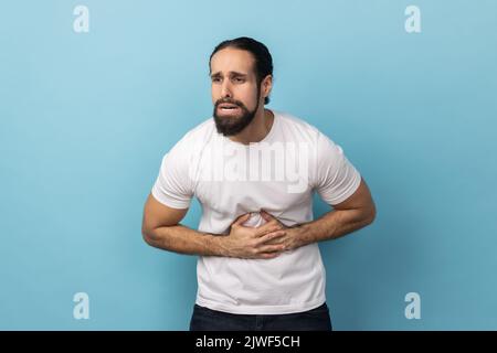 Portrait of ill sick man with beard wearing white T-shirt grimacing and suffering from pain in belly, severe abdominal distress Indoor studio shot isolated on blue background. Stock Photo