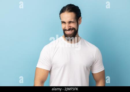 Portrait of cheerful man with beard wearing white T-shirt being in good mood, smiling broadly and winking at camera with toothy smile. Indoor studio shot isolated on blue background. Stock Photo