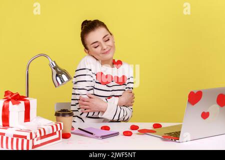 Woman covered with heart sticks sitting at workplace, hugging herself and smiling, feeling comfortable and fulfilled, egoistic person. Indoor studio studio shot isolated on yellow background. Stock Photo