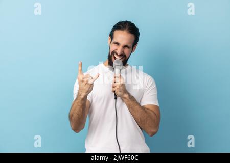 Portrait of excited man with beard in white T-shirt loudly singing song holding microphone in hand, having fun resting in karaoke, showing rock sign. Indoor studio shot isolated on blue background. Stock Photo