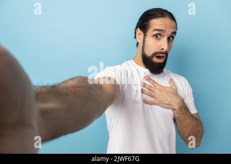 Portrait of scared shocked man with beard wearing white T-shirt looking at camera open mouth, keeps hand on chest, POV, point of view of photo. Indoor studio shot isolated on blue background. Stock Photo