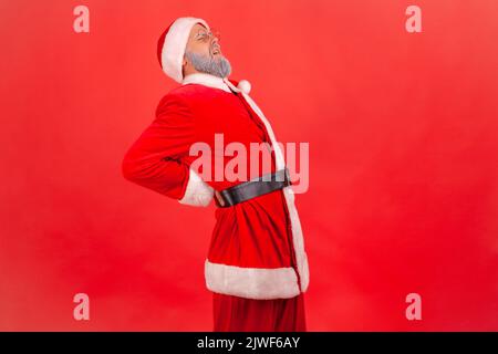 Sick elderly man with gray beard wearing santa claus costume feeling sharp pain in kidneys, back injury, pinched sciatic nerve, suffering. Indoor studio shot isolated on red background. Stock Photo