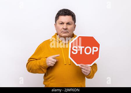 Portrait of middle aged strict bossy man pointing at Stop road traffic sign as symbol of prohibition, wearing urban style hoodie. Indoor studio shot isolated on white background. Stock Photo