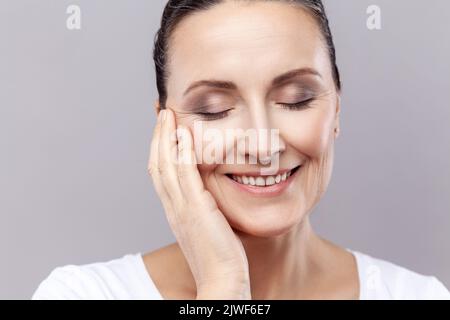 Closeup portrait of middle aged woman gently touching face with one hand and smiling, having healthy pure skin, female enjoying spa facial treatment. Indoor studio shot isolated on gray background. Stock Photo