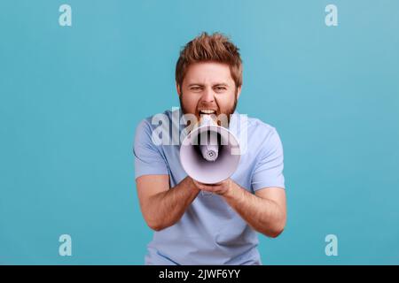Portrait of bearded man holding megaphone near mouth, loudly speaking, screaming, making announcement, paying attention at social problems. Indoor studio shot isolated on blue background. Stock Photo