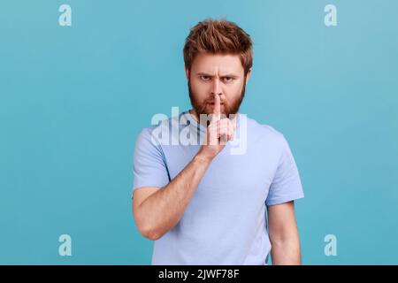 Portrait of bearded man holding finger near lips, showing shh gesture asking to keep silence and be quite, privacy, looking at camera. Indoor studio shot isolated on blue background. Stock Photo