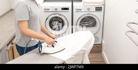 Woman ironing white clothes on board in laundry room with washing machine on background Stock Photo