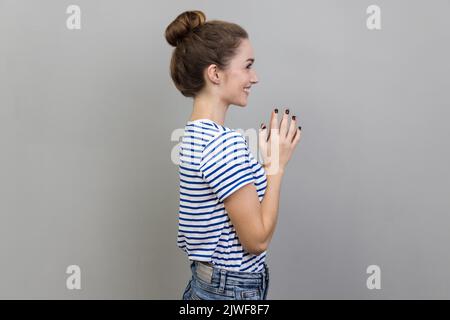 Side view portrait of cunning devious woman wearing striped T-shirt clasping hands and smirking mysteriously, scheming cheats, evil prank. Indoor studio shot isolated on gray background. Stock Photo