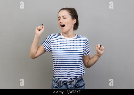 Portrait of tired sleepy woman wearing striped T-shirt yawning and raising hands up, feeling fatigued, standing with close eye. Indoor studio shot isolated on gray background. Stock Photo