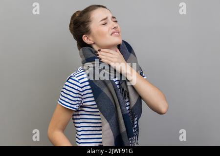 Sick woman in T-shirt and wrapped in scarf, touches neck, suffers from sore throat, cant breath well, chokes and smirks face, feels discomfort. Indoor studio shot isolated on gray background. Stock Photo