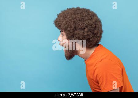 Side view of amazed man with Afro hairstyle wearing orange T-shirt standing with mouth open in surprise, has shocked expression, hears unbelievable news. Indoor studio shot isolated on blue background Stock Photo
