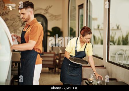 Waitress collecting dirty glassware from tablet when barista cleaning counter Stock Photo