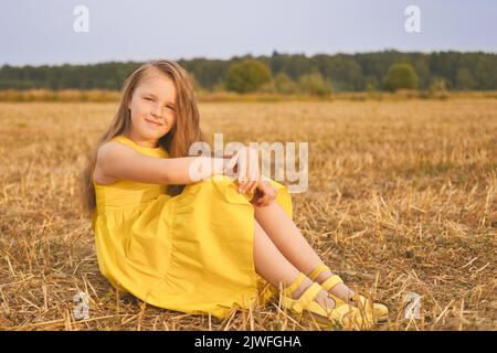 Cute little girl in yellow dress sits in the field hugging her knees. Beautiful girl with long hair outdoors. Stock Photo