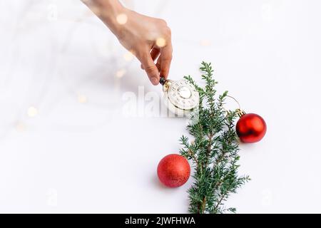 Woman's hand decorates Christmas tree branch with red round toys and New Year's clock on white background. Glowing yellow bokeh. Copy space for text, Stock Photo