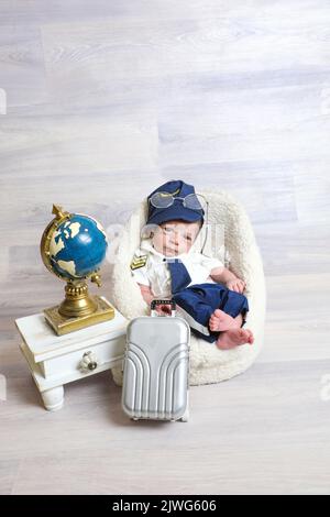 toddler boy in cute pilot costume lying at soft tiny chair little children props travel suitcase globe statue on table Stock Photo