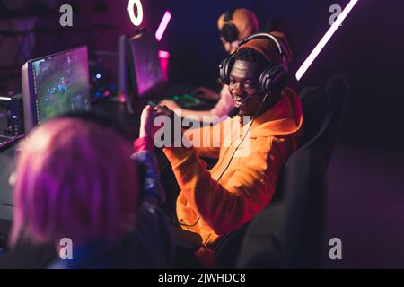 Three teenagers wearing colorful clothes playing online game using professional gaming setup celebrating victory in tournament with fist bumps. Color neon lights. Indoor shot. High quality photo Stock Photo