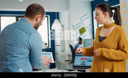 Man in startup office zoomin in digital tablet using touchscreen presenting sales charts to woman colleague standing next to desk. Team of two coworkers working on group marketing project. Stock Photo